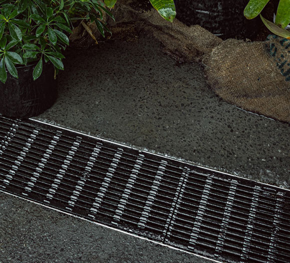 A black metal grate lines a textured surface, surrounded by potted plants and a burlap sack, creating a lush, outdoor setting.
