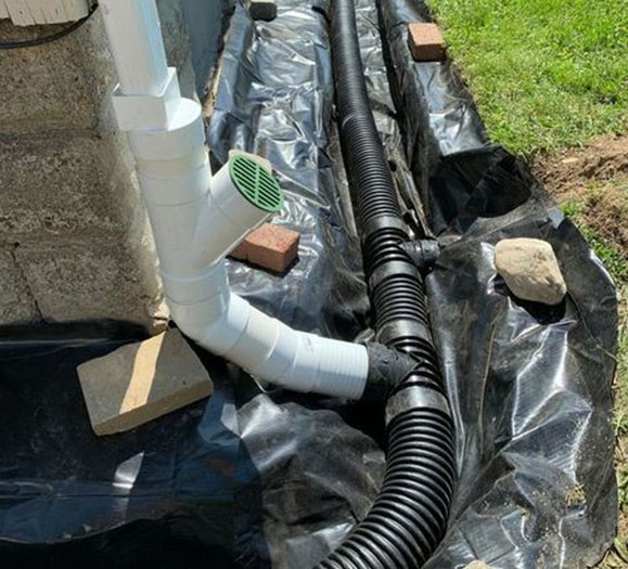 A drainpipe system made of white PVC and black flexible tubing is installed on a gravel surface, covered with black plastic sheeting.