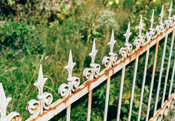 Close-up of a weathered white wrought iron fence with decorative finials, surrounded by lush green grass and wildflowers.